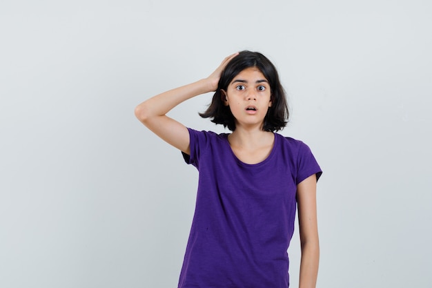 Little girl in t-shirt holding hand on head and looking puzzled ,