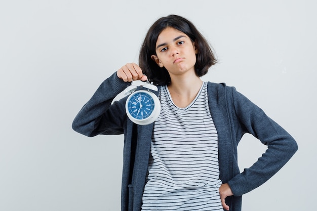 Little girl in t-shirt, jacket holding alarm clock and looking sad ,