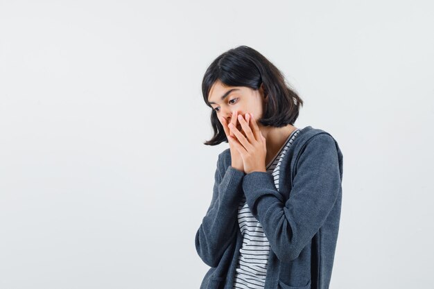 Little girl in t-shirt, jacket holding hands on mouth and looking surprised