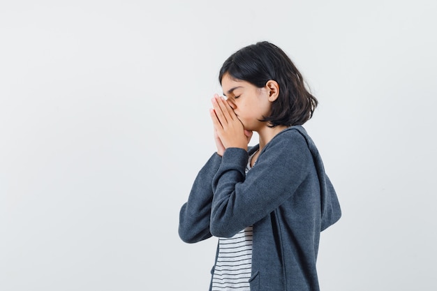 Little girl in t-shirt, jacket holding hands in praying gesture and looking hopeful .