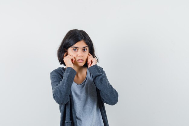 Little girl in t-shirt, jacket plugging ears with fingers and looking puzzled