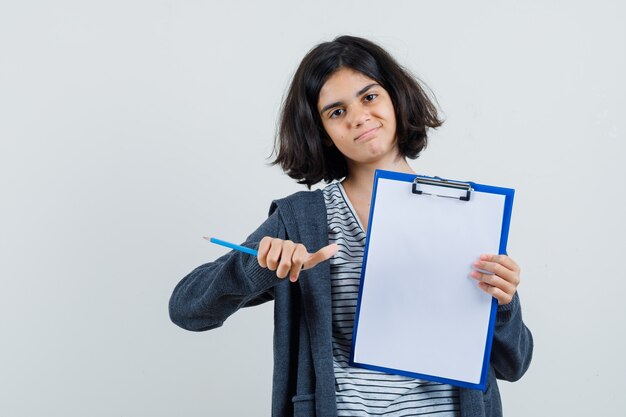 Little girl in t-shirt, jacket pointing at clipboard, holding pencil and looking confident ,