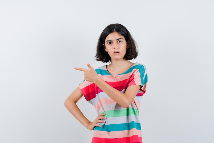 मुफ़्त फोटो टी-शर्ट, जींस में छोटी लड़की कमर पर हाथ रखकर बाएँ ओर इशारा कर रही है और हैरान दिख रही है, सामने का दृश्य।