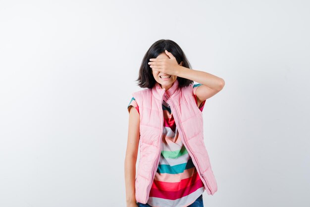 Little girl in t-shirt, puffer vest, jeans covering eyes with hand and looking scared , front view.