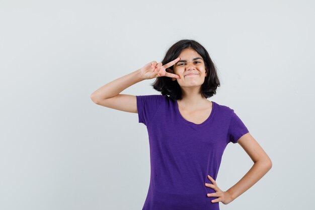 Little girl in t-shirt showing v-sign near eye and looking amused ,