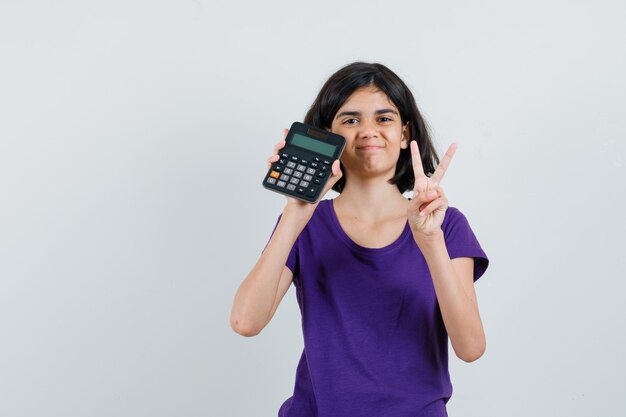 Little girl in t-shirt showing victory gesture, holding calculator and looking merry ,