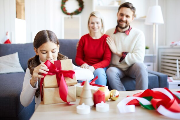Маленькая девочка разворачивает рождественский подарок, пока ее родители счастливо смотрят на нее.