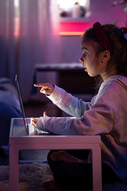 Free photo a little girl uses a laptop while sitting in a room with neon lighting