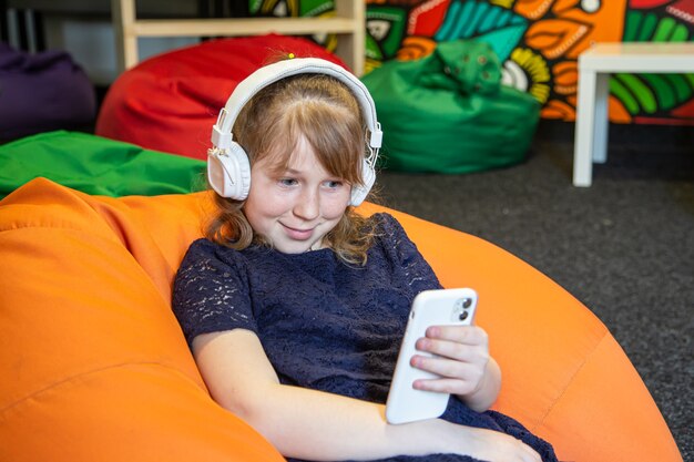 Little girl uses phone and listens to music with headphones while sitting on bag chair.