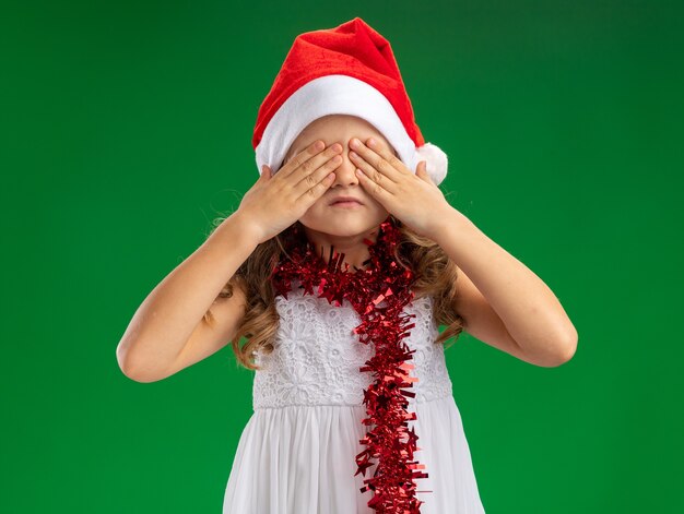 Little girl wearing christmas hat with garland on neck covered eyes with hands isolated on green wall