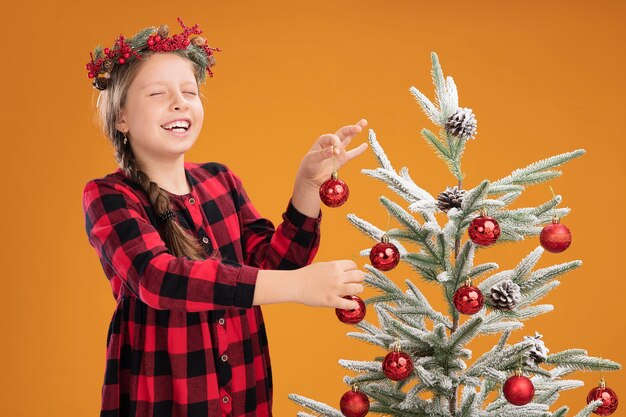 Little girl wearing christmas wreath in checked dress decorating christmas tree happy and cheerful standing over orange wall