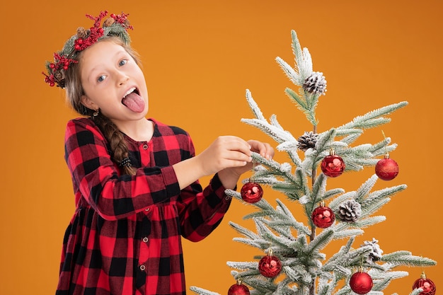 Little girl wearing christmas wreath in checked dress   decorating  christmas tree  happy and joyful sticking out tongue over orange background