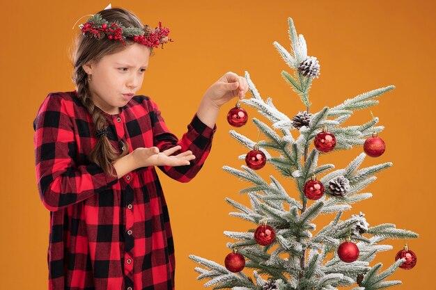 Little girl wearing christmas wreath in checked dress decorating christmas tree  looking confused and very anxious standing over orange background