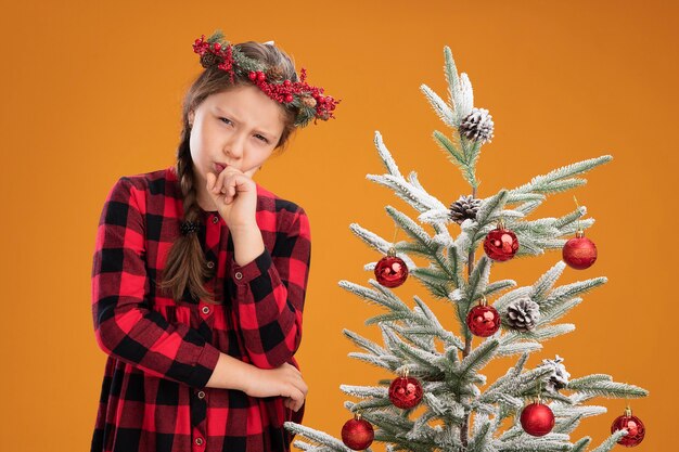 オレンジ色の壁の上のクリスマスツリーの横に立っている真面目な顔で考えているあごに手でチェックドレスでクリスマスリースを着ている少女