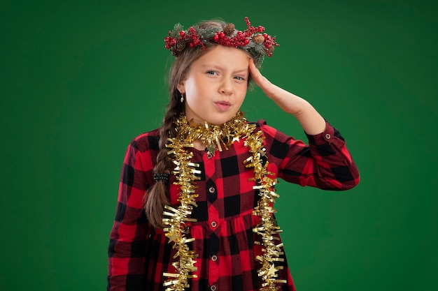 Free photo little girl wearing christmas wreath in checked dress  with tinsel around neck confused with hand on her head for mistake