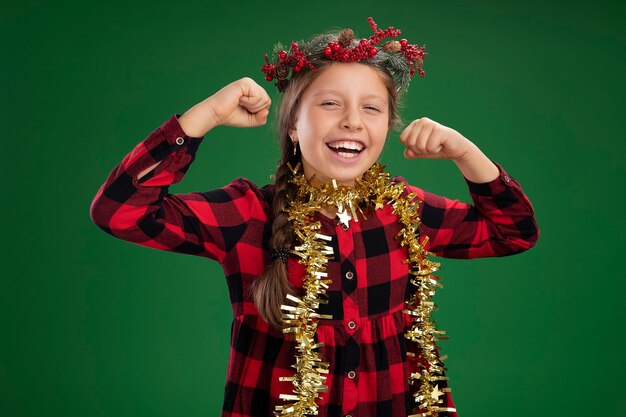 首の周りに見掛け倒しの幸せで興奮した握りこぶしでチェックのドレスでクリスマスリースを着ている少女