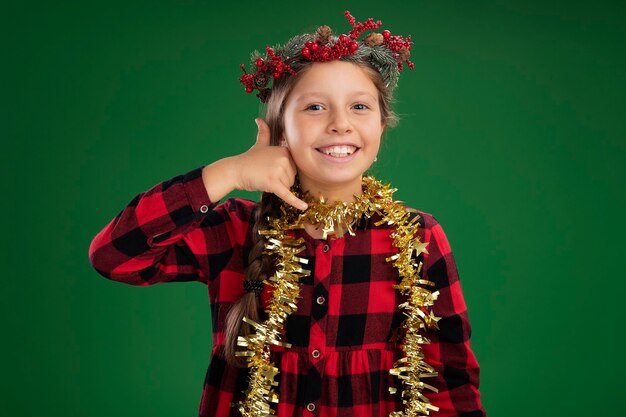 Маленькая девочка в рождественском венке в клетчатом платье с мишурой на шее счастливая и позитивная, делая жест «Позвони мне», стоя над зеленой стеной