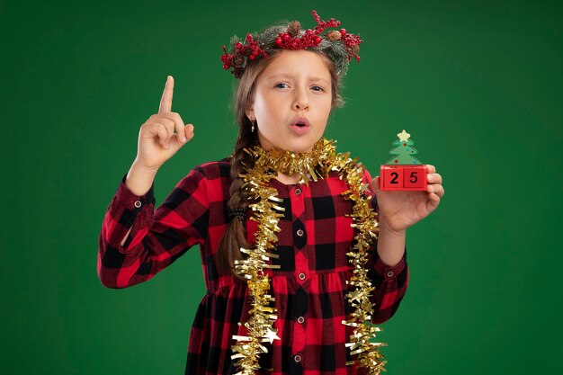 Маленькая девочка в рождественском венке в клетчатом платье с мишурой на шее держит игрушечные кубики с удивленным свиданием, показывая указательный палец