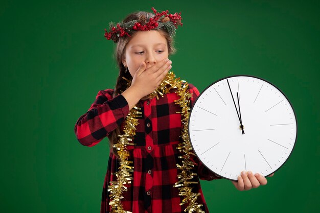 Little girl wearing christmas wreath in checked dress  with tinsel around neck holding wall clock looking at it being shocked covering mouth with hand standing over green wall