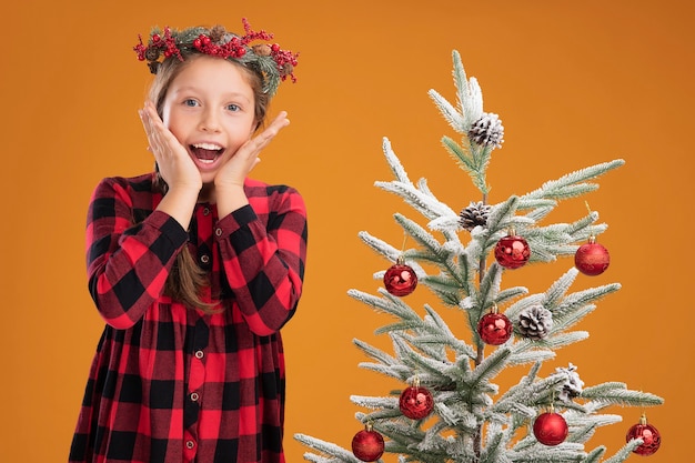 Free photo little girl wearing christmas wreath in checked shirt  happy and excited  standing next to a christmas tree over orange wall