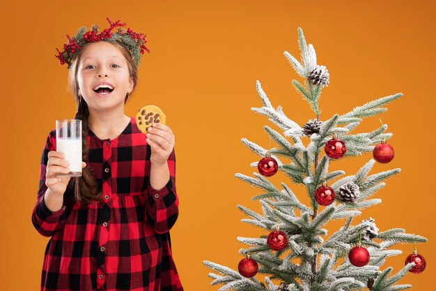 オレンジ色の壁の上のクリスマスツリーの横に立って幸せで陽気なミルクとクッキーのガラスを保持しているチェックシャツにクリスマスリースを着ている少女