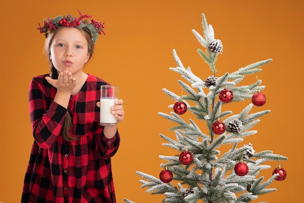 Bambina che indossa ghirlanda di natale in camicia a quadri con in mano un bicchiere di latte felice e positiva che soffia un bacio in piedi accanto a un albero di natale sul muro arancione