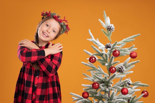 Little girl wearing christmas wreath in checked shirt holding hands on chest happy and positive feeling thankful standing next to a christmas tree  over orange wall