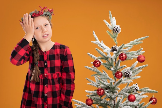 Little girl wearing christmas wreath in checked shirt looking confused with hand on her head standing next to a christmas tree  over orange wall