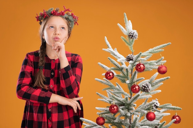 체크 셔츠에 크리스마스 화 환을 착용하는 어린 소녀 오렌지 벽 위에 크리스마스 트리 옆에 의아해 서 찾고