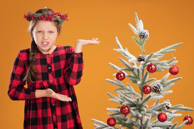 Little girl wearing christmas wreath in checked shirt standing next to a christmas tree looking confused raising arms in displeasure and indignation  over orange wall