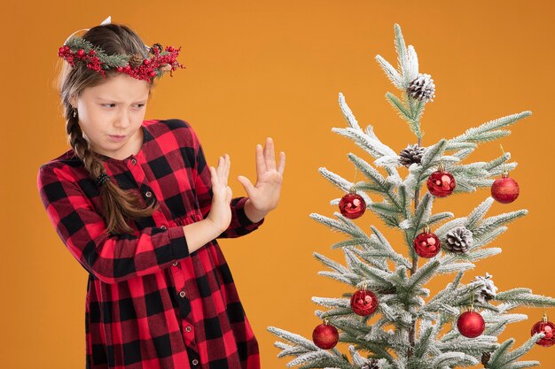 Little girl wearing christmas wreath in checked shirt standing next to a christmas tree looking at it with disgusted expression holding hands out making defense gesture over orange wall