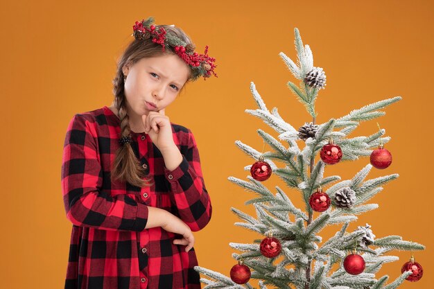 Маленькая девочка в рождественском венке в клетчатой рубашке, положив руку на подбородок, думает с серьезным лицом, стоит рядом с елкой над оранжевой стеной