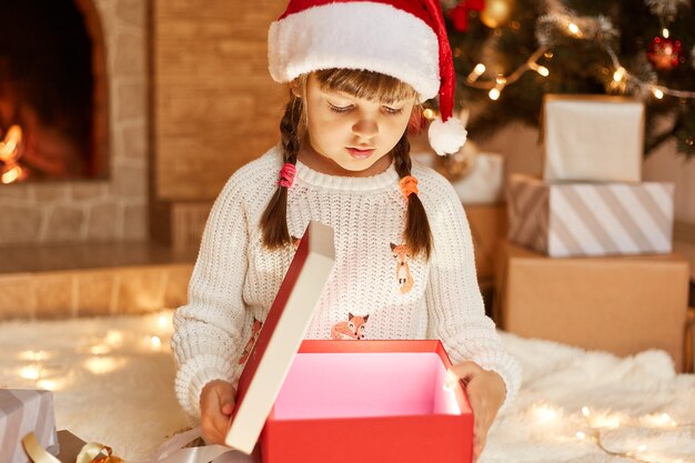 白いセーターとサンタクロースの帽子をかぶった少女は、暖炉とクリスマスツリーのあるお祭りの部屋でポーズをとって、中に何かが光っているプレゼントボックスを開きます。