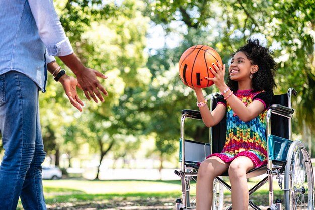 A little girl in a wheelchair having fun with her father while playing basketball together at park