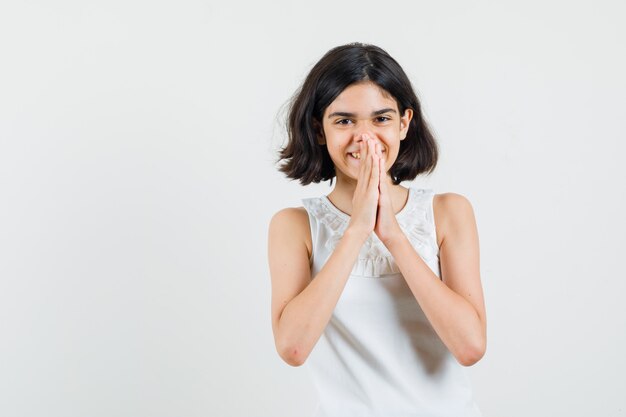 Little girl in white blouse holding hands in praying gesture and looking cheerful , front view.