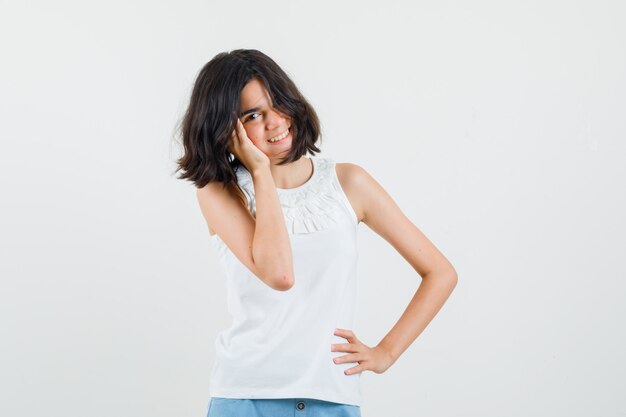 Little girl in white blouse, shorts holding hand on cheek and looking cute , front view.