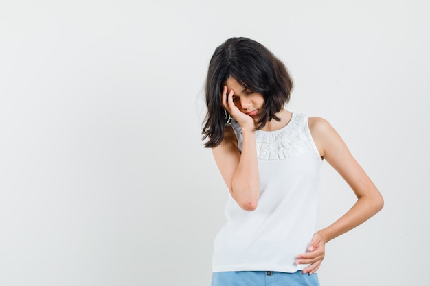 Free photo little girl in white blouse, shorts holding hand on face and looking exhausted , front view.