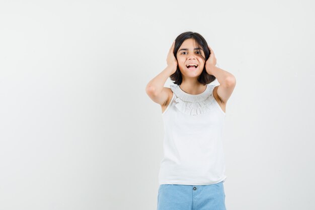 Little girl in white blouse, shorts holding hands on ears , front view.