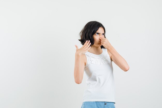 Little girl in white blouse, shorts pinching nose due to bad smell and looking disgusted , front view.