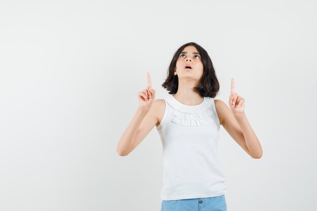 Little girl in white blouse, shorts pointing up and looking amazed , front view.