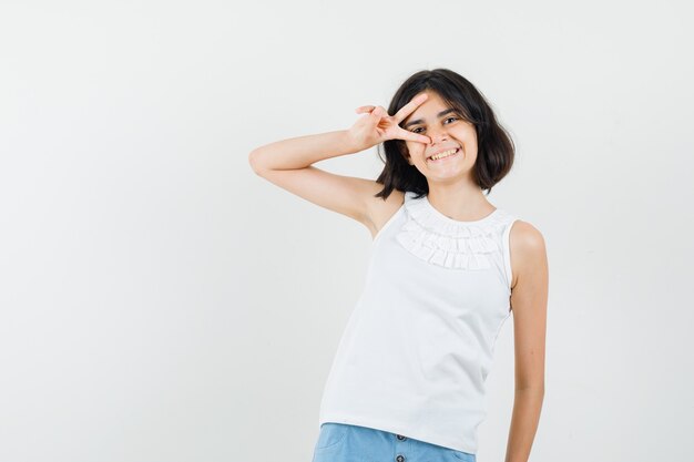 Little girl in white blouse, shorts showing v-sign near eye and looking pretty , front view.