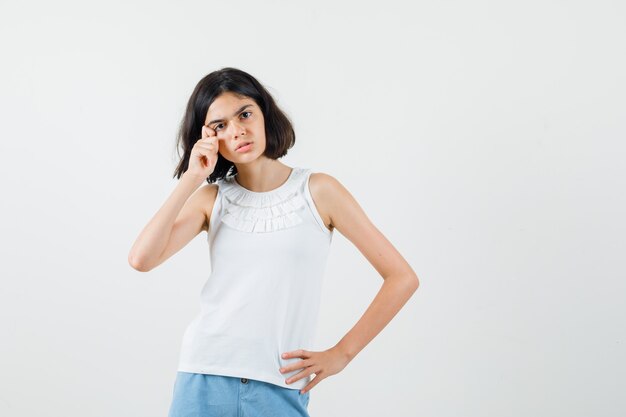 Little girl in white blouse, shorts wiping tears by fingers and looking sad , front view.