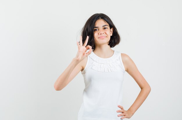 Little girl in white blouse showing ok sign and looking confident , front view.