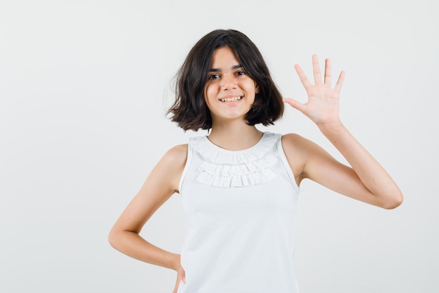 Little girl in white blouse waving hand to say hello or goodbye and looking merry , front view.