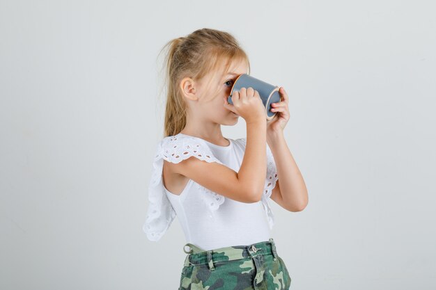 Free photo little girl in white t-shirt, skirt drinking cup of tea and looking thirsty