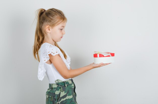 Little girl in white t-shirt, skirt giving someone gift box and looking careful