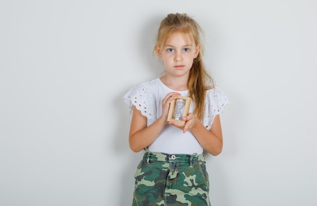 Little girl in white t-shirt, skirt holding hourglass and looking careful