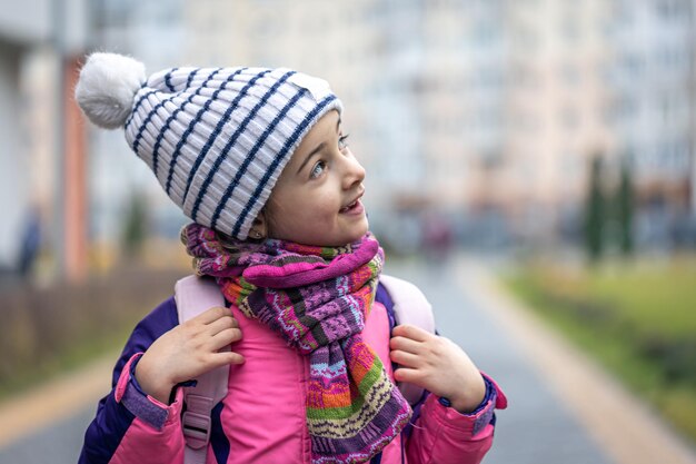 Little girl with a backpack in a jacket and a hat near the school copy space