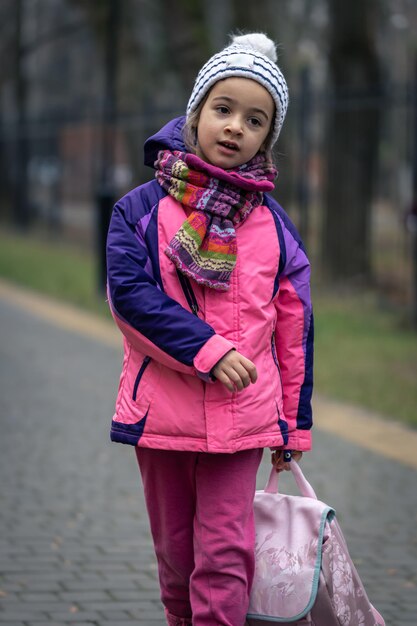 Little girl with a backpack in a jacket and a hat near the school