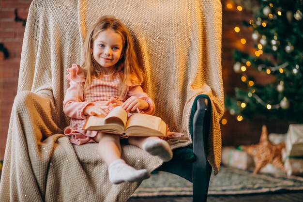 Little girl with book sitting in chair by christmas tree